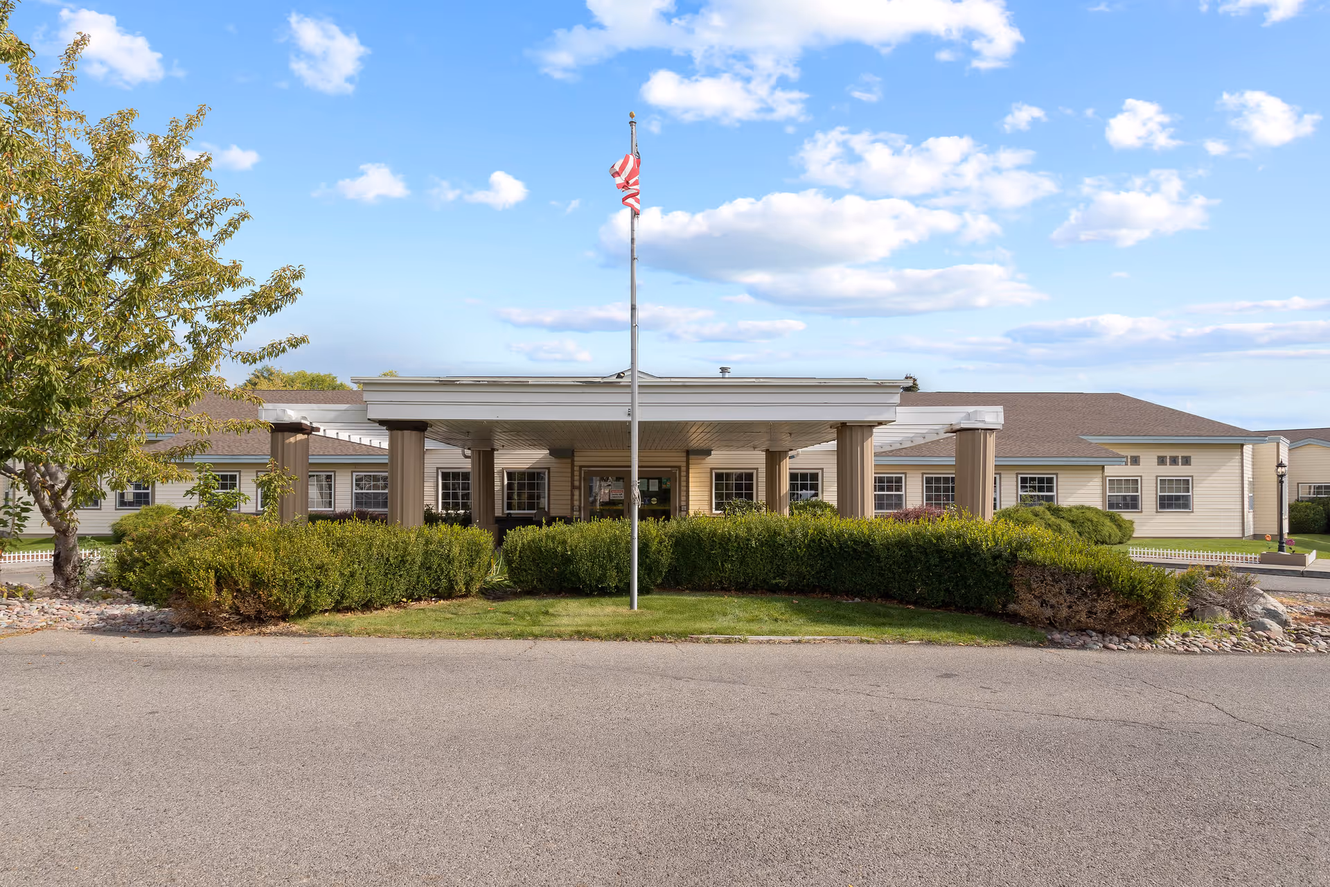 Front entrance of Sullivan Park Care Center with a covered portico, flagpole, and landscaped shrubs under a blue sky.