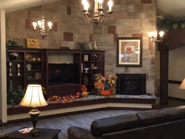 A cozy living room area with a stone accent wall featuring a built-in entertainment center with a TV and shelves decorated with vases, books, and flowers. There is a fireplace below a framed floral artwork. The room is softly lit by multiple chandeliers and a table lamp on a side table next to a dark leather sofa.