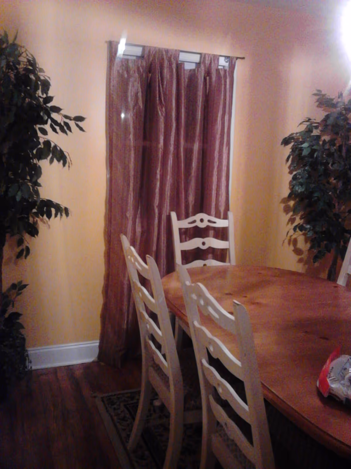 Dining room with a wooden table and white ladder-back chairs, flanked by potted plants and a window with burgundy curtains.