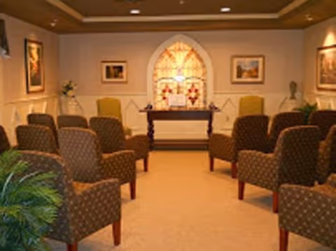 A small chapel or meditation room with rows of patterned upholstered chairs facing a wooden altar table. Behind the altar is a stained glass window with a cross design. The room has soft lighting, framed pictures on the walls, and plants in the corners.