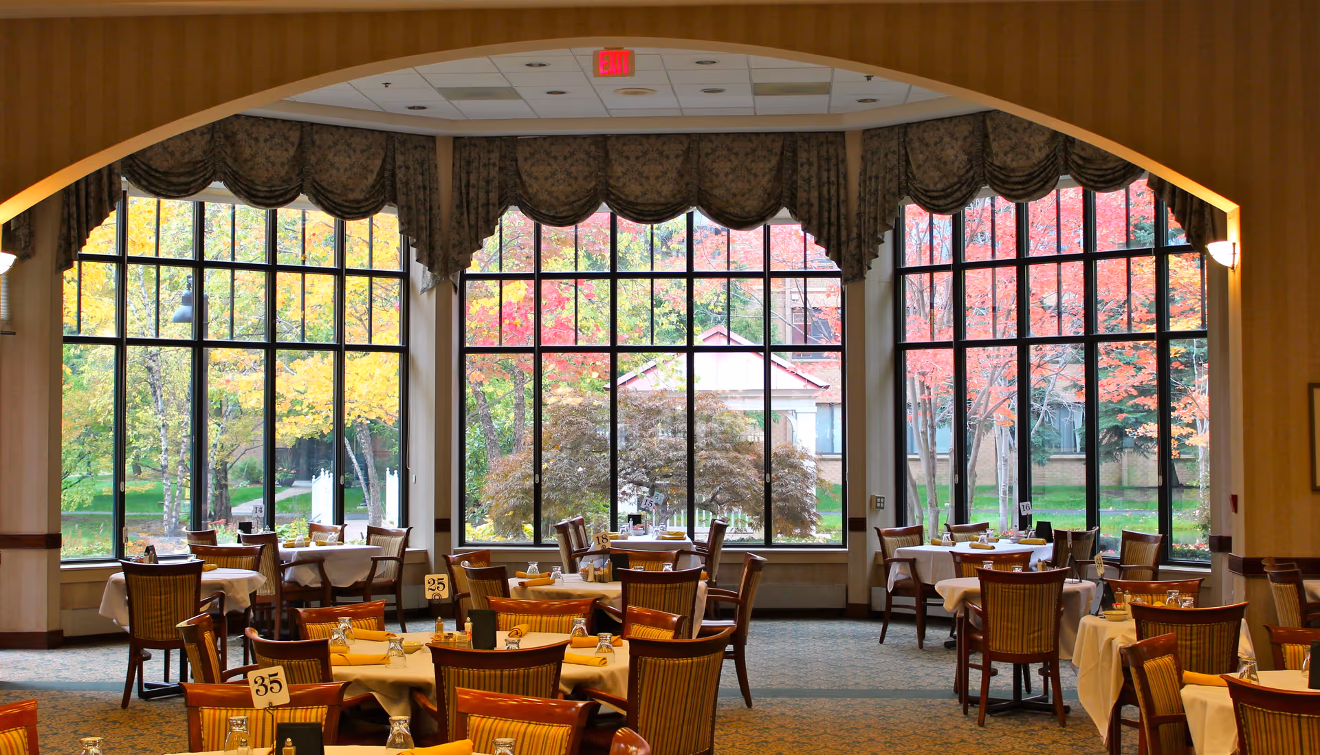 A dining room with multiple tables covered in white tablecloths, each set with glasses and napkins. Large windows with decorative valances provide a view of colorful autumn trees outside.