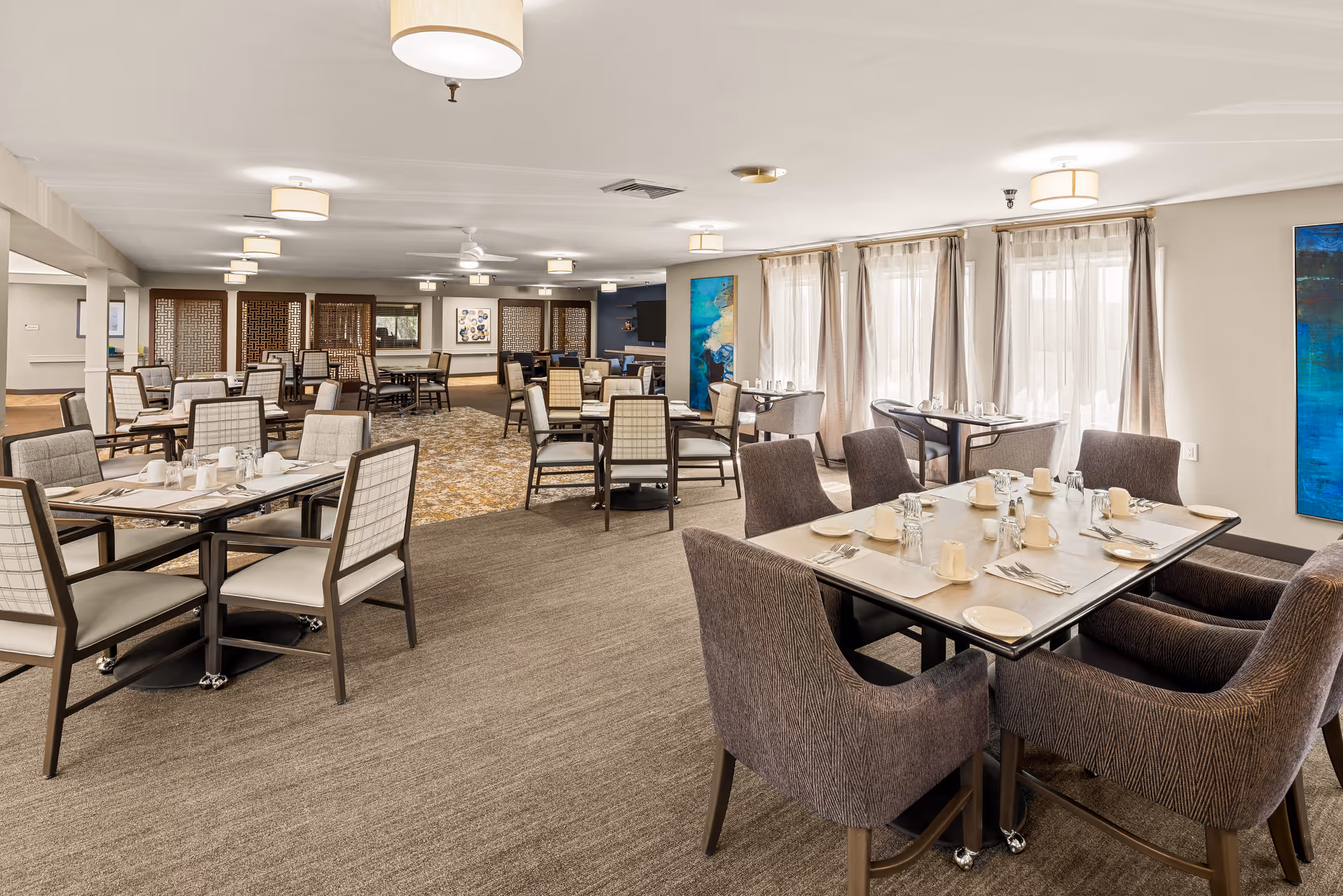 A spacious dining room in a senior living community with multiple tables and chairs arranged neatly. The tables are set with plates, cups, and silverware. Large windows with sheer curtains allow natural light to fill the room. The walls are decorated with colorful abstract paintings, and ceiling lights provide additional illumination.