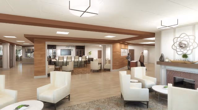 A spacious and modern senior living facility common area with light-colored armchairs arranged around small round tables. The room features wooden paneling on the walls, a fireplace with decorative items on the mantel, and a dining area with a long table and chairs in the background. Two people are standing and conversing near the dining area.