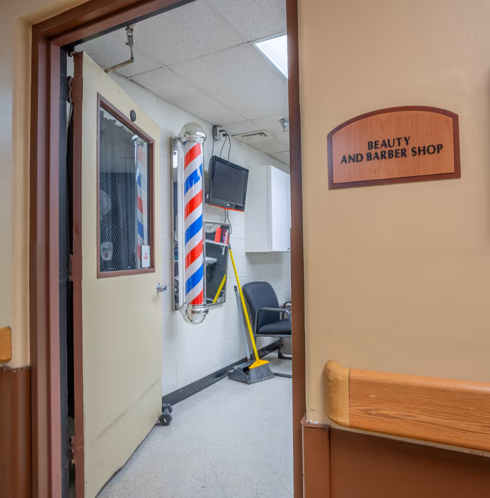 View through an open door into a beauty and barber shop inside a facility. Visible inside are a traditional barber pole with red, white, and blue stripes, a black chair, a broom and dustpan, a wall-mounted TV, and a mirror. The door has a window with wire mesh, and a wooden sign on the wall outside the door reads 'Beauty and Barber Shop'.