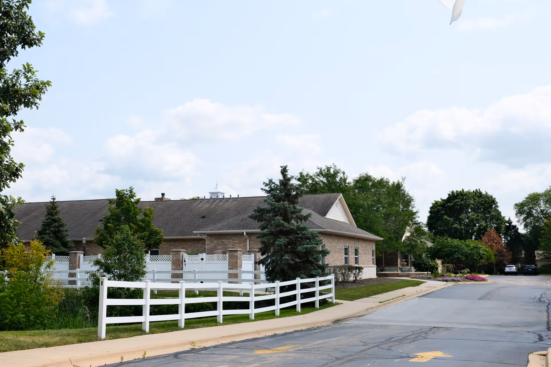 Exterior view of a single-story brick building with a gray roof, surrounded by trees and a white fence. The scene includes a paved driveway and a partly cloudy sky.