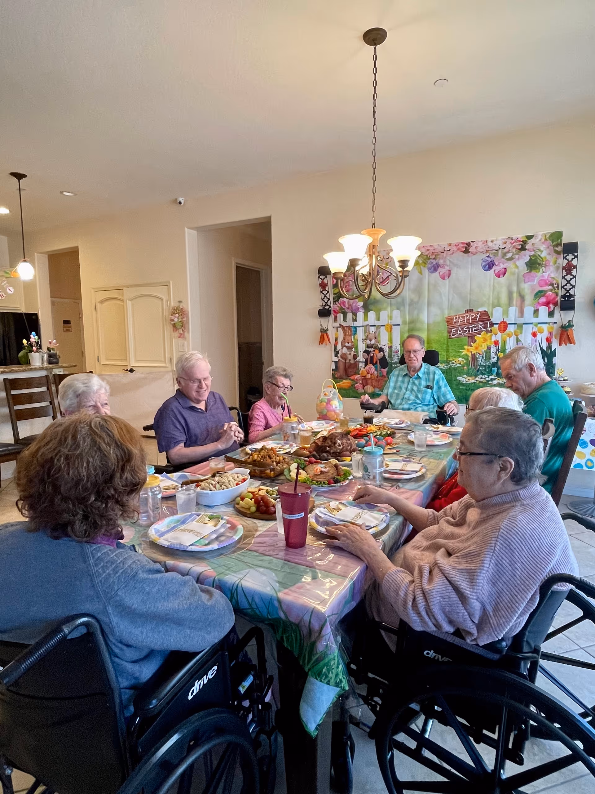 A group of elderly people, some in wheelchairs, sitting around a dining table set with plates of food and drinks in a well-lit room decorated with an Easter-themed backdrop and a chandelier overhead.