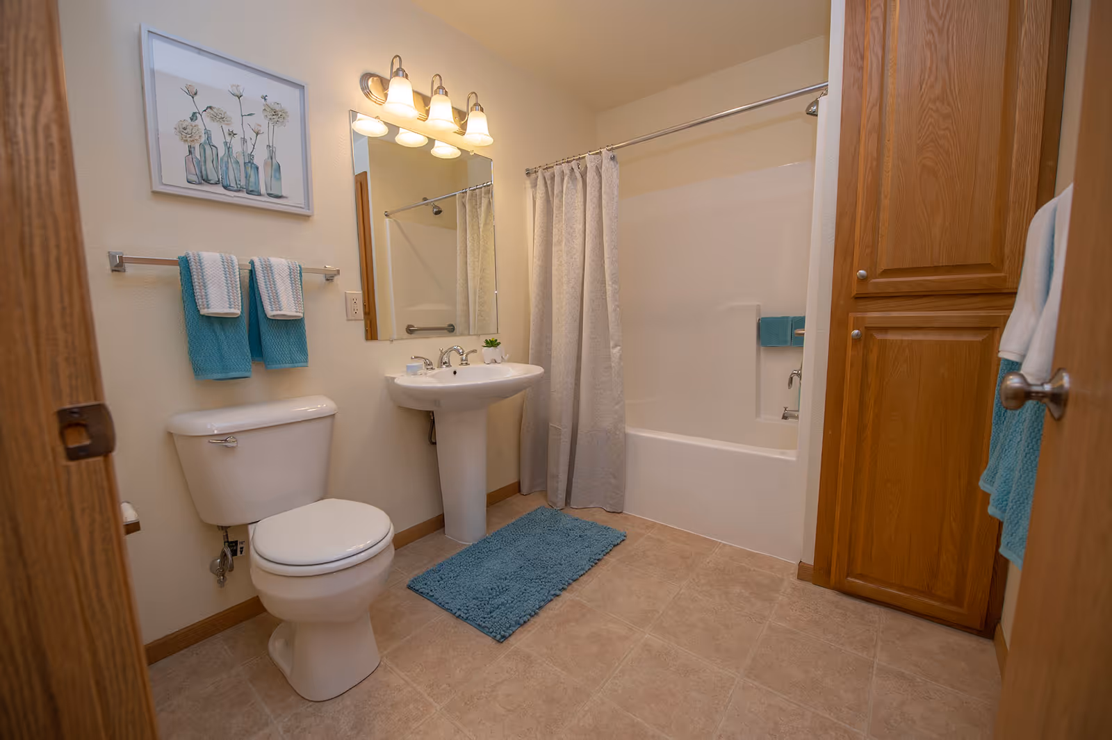 Clean bathroom interior featuring a toilet, pedestal sink, bathtub with shower curtain, wood storage cabinet and teal towels.