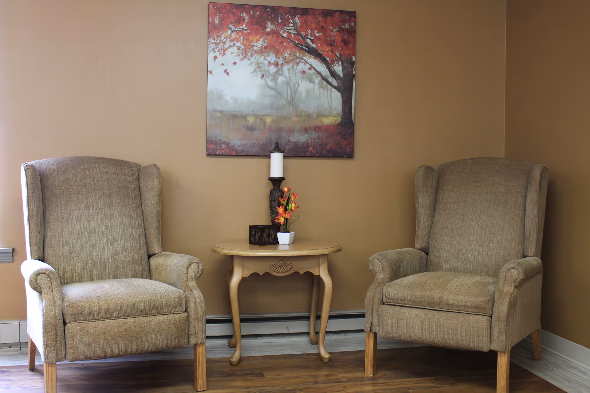 Two beige upholstered armchairs with wooden legs are positioned on either side of a small wooden table. On the table, there is a decorative candle holder with a white candle, a small potted plant with orange flowers, and a small dark box. Above the table hangs a painting of a tree with red leaves in a misty landscape. The walls are painted light brown and the floor is wooden.