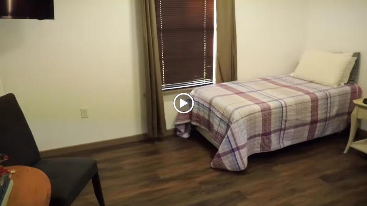 A small bedroom with a single bed covered in a plaid quilt, a pillow, a window with closed blinds and curtains, a dark upholstered chair, a small round wooden table with a decorative item, and a wall-mounted TV.