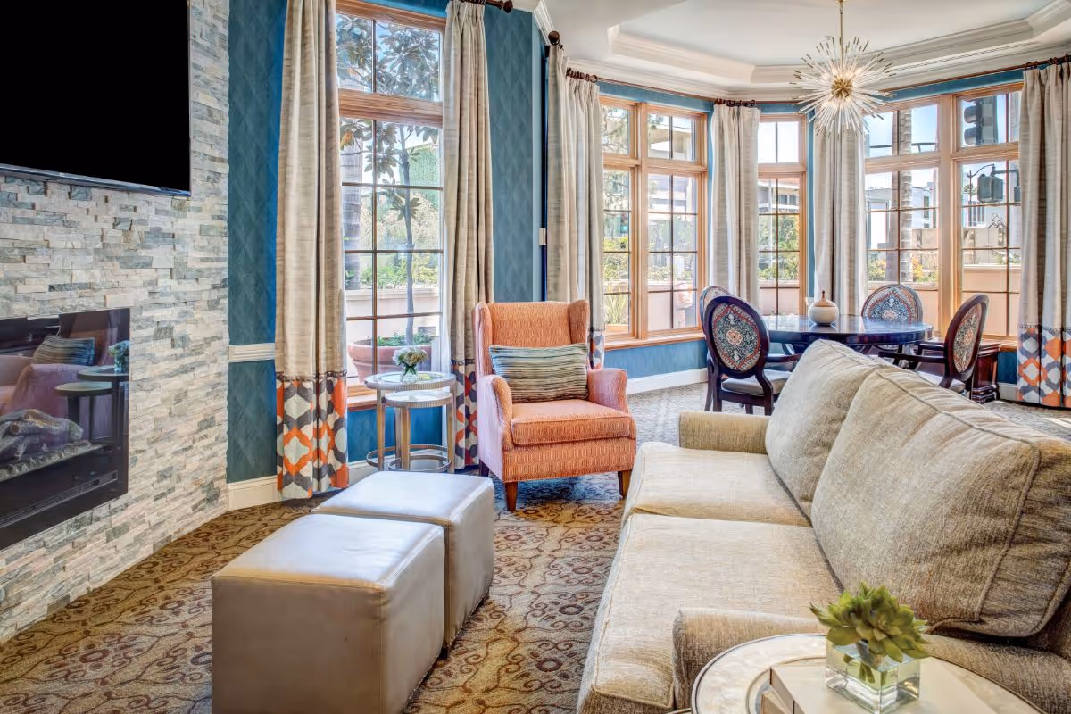 A bright and cozy living room area with a beige sofa, two gray ottomans, and an orange armchair with a striped pillow. The room features large windows with beige and patterned curtains, allowing natural light to fill the space. There is a stone accent wall with a mounted flat-screen TV and a modern fireplace below it. In the background, a round dining table with four upholstered chairs is visible under a decorative chandelier.