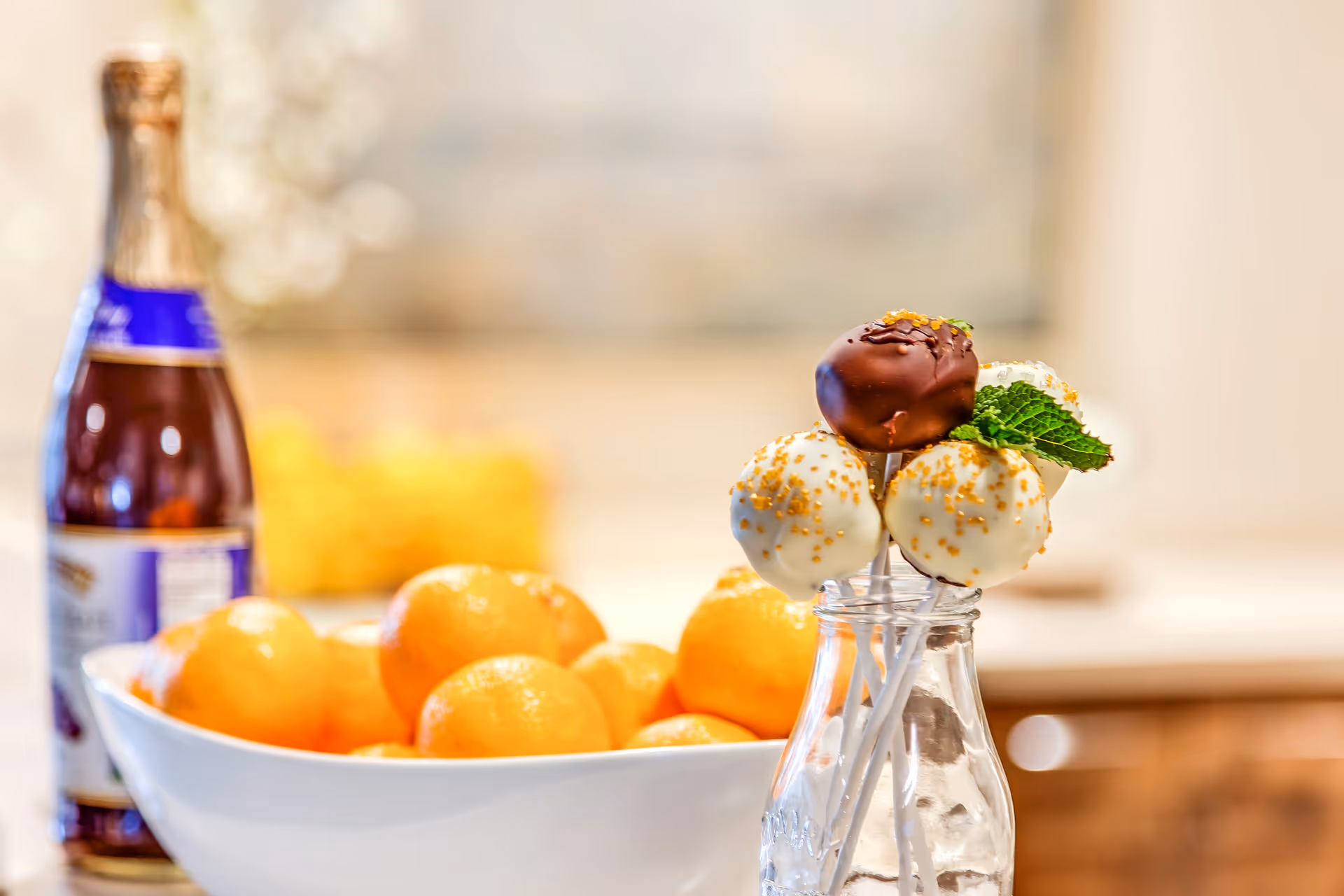 A kitchen counter with a bowl of oranges, a bottle, and chocolate-covered cake pops in a small glass jar.