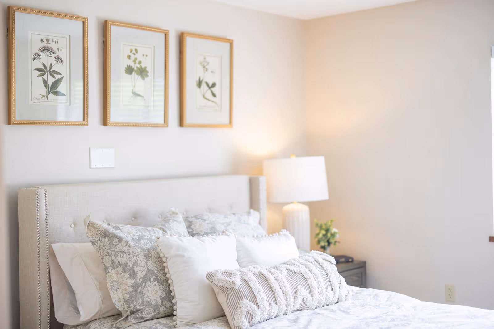 Light, airy bedroom with a tufted beige headboard, layered decorative pillows, three framed botanical prints above the bed, and a bedside lamp.