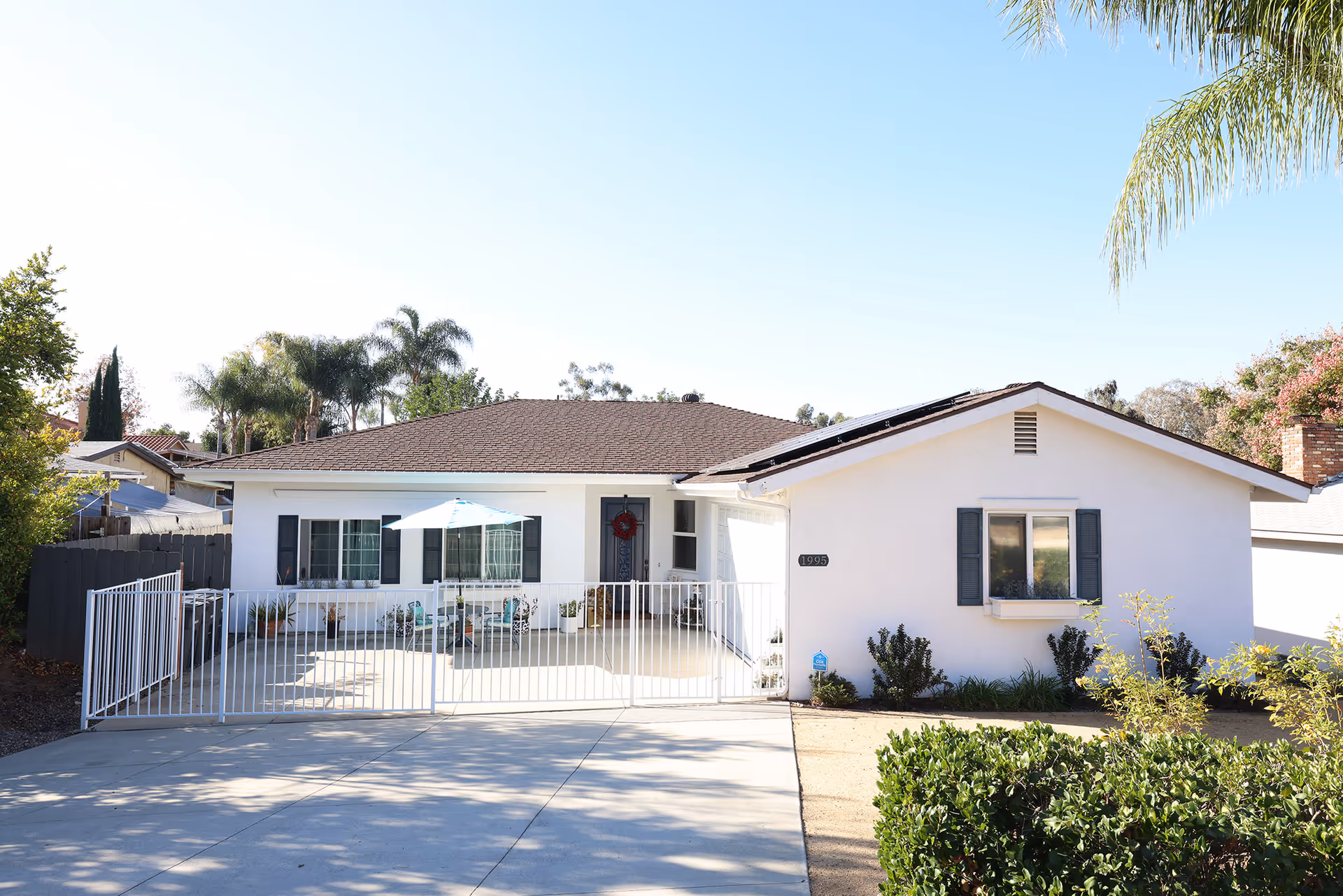 Front exterior view of a single-story white house with a brown roof, a gated front patio with outdoor furniture and an umbrella, surrounded by greenery and palm trees under a clear sky.