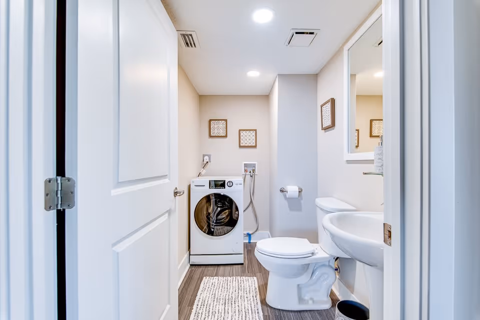 Bathroom featuring a toilet, pedestal sink with mirror, and a front-loading washing machine in a compact laundry-bath combo.