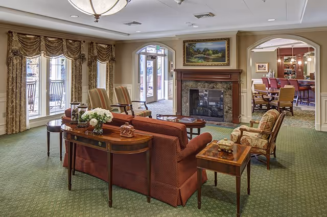 A cozy senior living facility common area with a red sofa, patterned armchairs, wooden side tables, and a fireplace with a landscape painting above it. Large windows with draped curtains allow natural light, and a dining area with chairs and tables is visible through an archway.