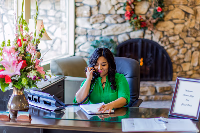 Reception desk with a woman on the phone, a vase of flowers, paperwork and a guest book sign in front of a stone fireplace.