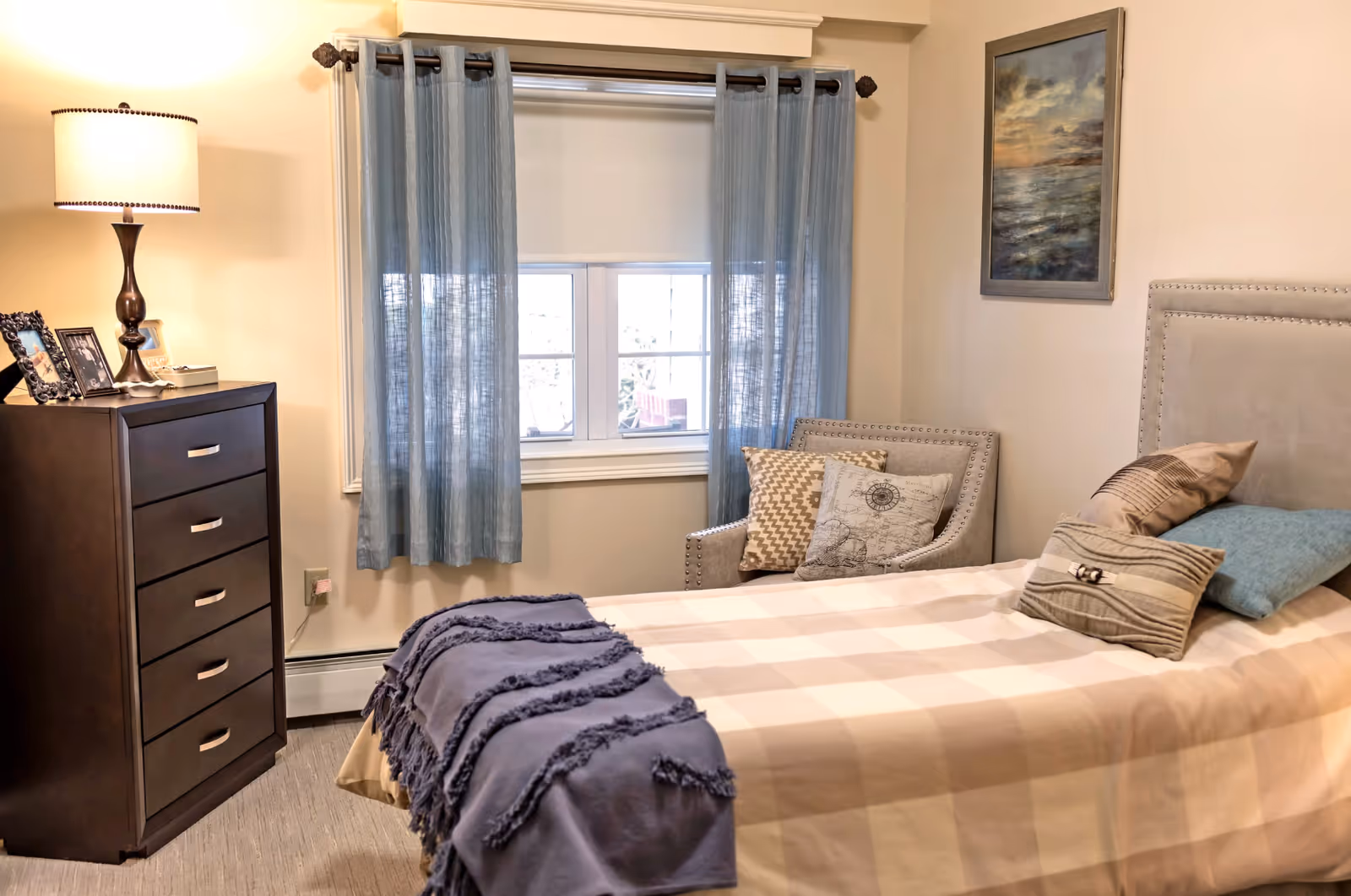 A cozy bedroom with a single bed covered in a beige checkered blanket and several decorative pillows. Next to the bed is a gray armchair with two patterned cushions. A dark wooden dresser with a lamp and framed photos sits against the wall. A window with blue curtains is centered on the wall behind the bed and chair. A framed painting hangs on the wall above the chair.