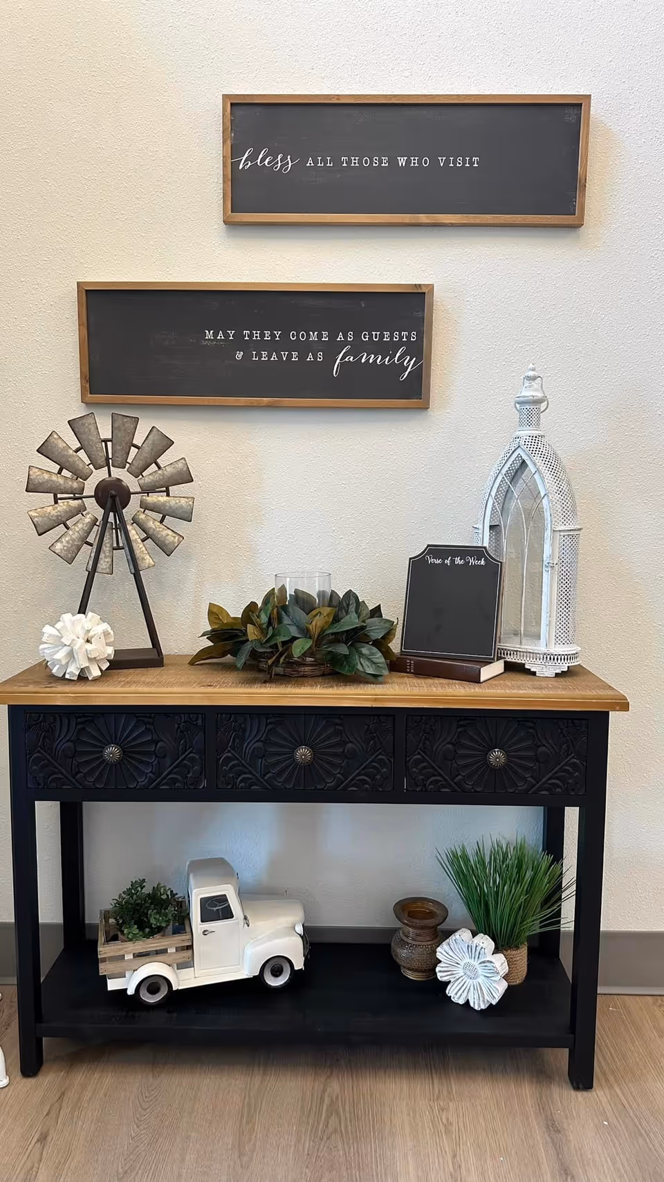 A decorative console table with three drawers and a wooden top against a beige wall. On the table are various decorative items including a small metal windmill, a white ornamental object, a green leafy centerpiece, a small chalkboard sign labeled 'Verse of the Week,' a book, and a white lantern. Below the table are a small white toy truck carrying plants, two small vases, a white flower decoration, and a potted grass plant. Above the table are two framed blackboards with white text that read 'bless ALL THOSE WHO VISIT' and 'MAY THEY COME AS GUESTS & LEAVE AS family'.