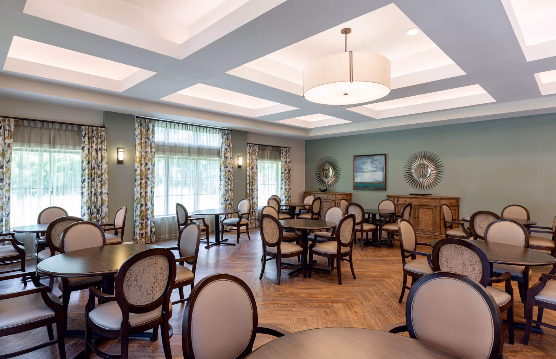 A spacious dining room with multiple round and oval dark wood tables surrounded by cushioned chairs with beige upholstery. The room features large windows with floral curtains, soft wall sconces, a coffered ceiling with recessed lighting, and two wooden sideboards against a green wall decorated with two sunburst mirrors and a framed painting.