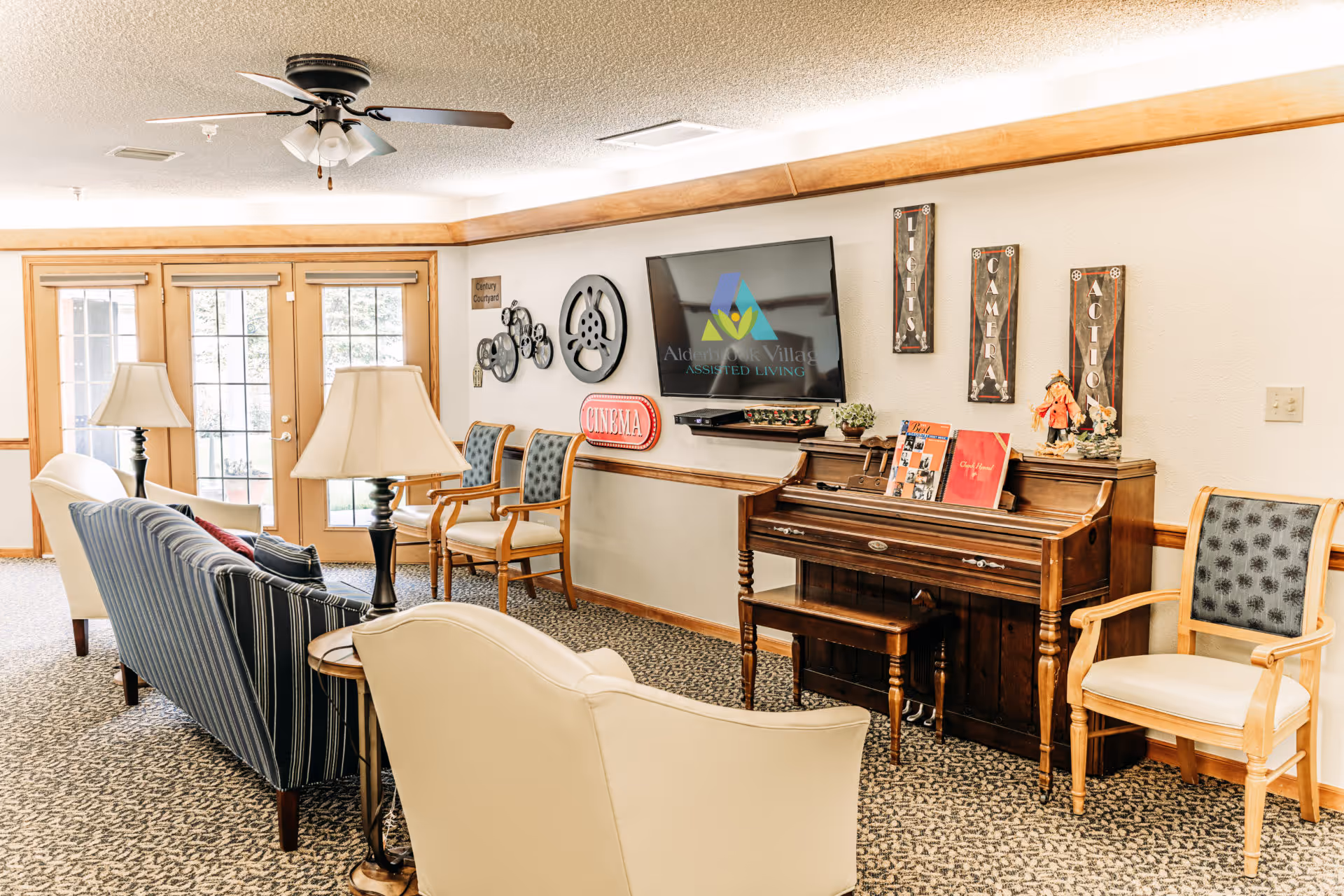 A cozy living room area in Alderbrook Village assisted living facility featuring a striped blue couch, cream armchairs, two table lamps, a ceiling fan, and a wooden piano with decorative items on top. A flat-screen TV mounted on the wall displays the Alderbrook Village logo. The wall is decorated with film-themed signs and artwork. French doors with windows lead to an outdoor courtyard.