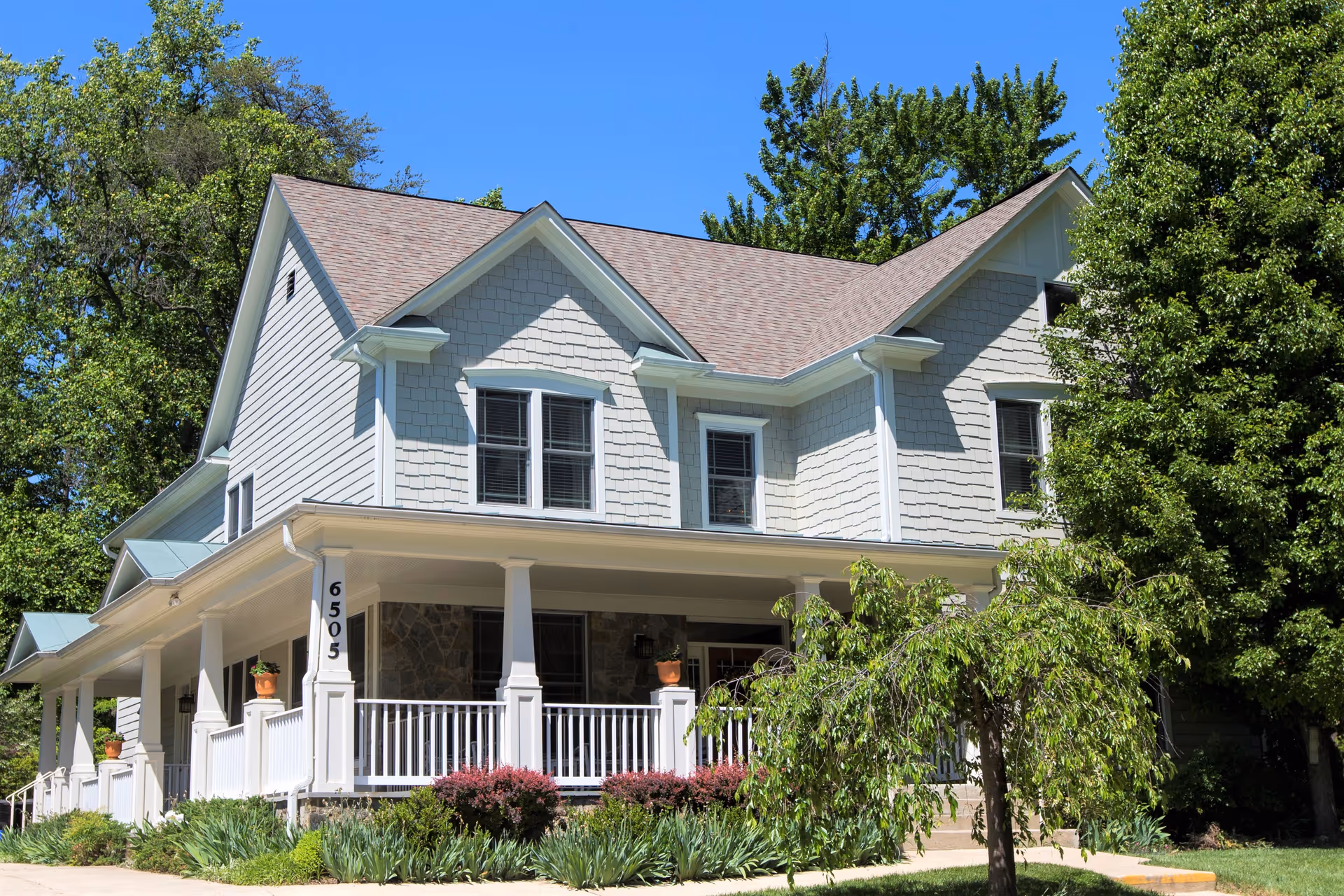 A large two-story house with light gray siding and a wrap-around porch with white railings. The house is surrounded by green trees and shrubs under a clear blue sky.