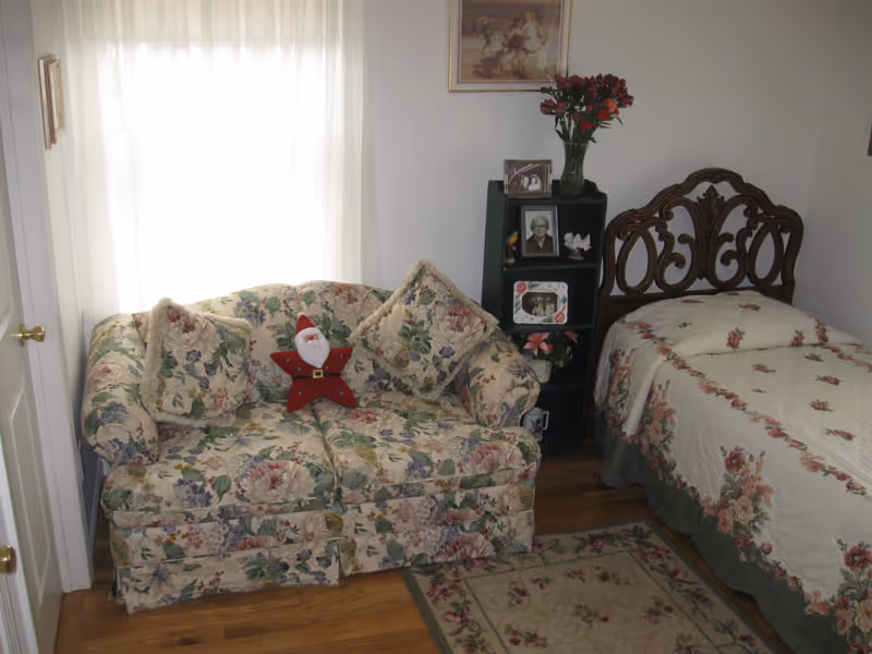 A cozy bedroom with a floral patterned loveseat and matching cushions, a single bed with a floral bedspread, a small black shelving unit holding framed photos and a vase with red flowers, and a floral rug on a wooden floor.