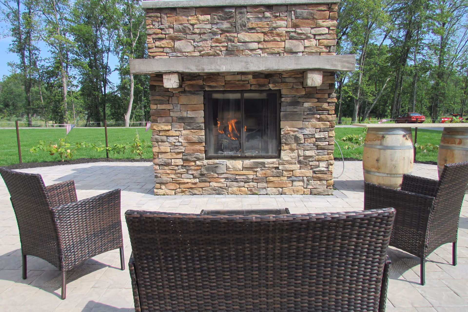 Outdoor patio area with a stone fireplace that has a fire burning inside. In front of the fireplace are three dark wicker chairs arranged around the fire. The patio is paved with stone tiles and surrounded by green grass and trees in the background. Two wooden barrels are visible on the right side of the fireplace.