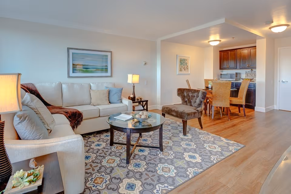 Open living area with a beige sofa, accent chair, glass coffee table on a patterned rug and an adjacent dining area and kitchenette.