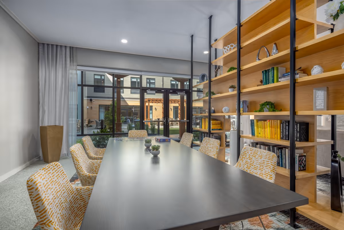 A modern meeting or dining room with a long black table surrounded by patterned yellow and white chairs. The room features a large wooden bookshelf filled with books and decorative items. Large glass doors and windows provide a view of an outdoor courtyard with seating and greenery.