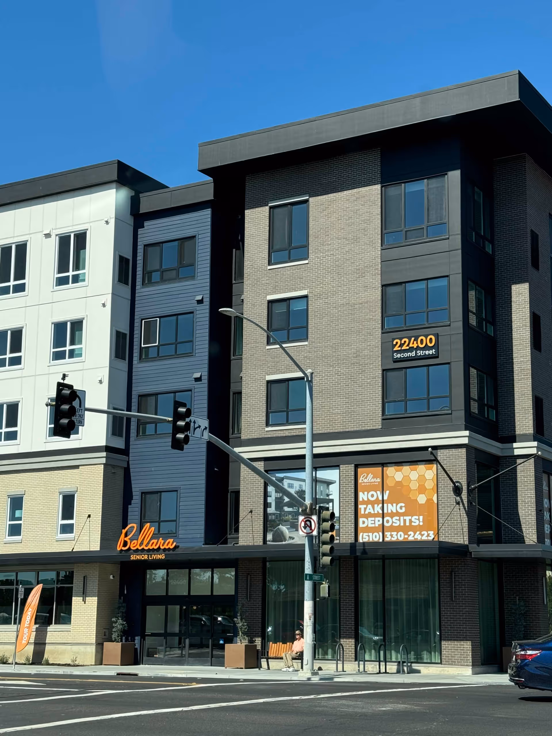 Exterior view of Bellara Senior Living facility, a modern multi-story building with large windows and a sign displaying the address 22400 Second Street. There is an orange sign on the building that reads 'Bellara Senior Living NOW TAKING DEPOSITS! (510) 330-2423'. A person is sitting on a bench near the entrance, and a traffic light is visible at the intersection.