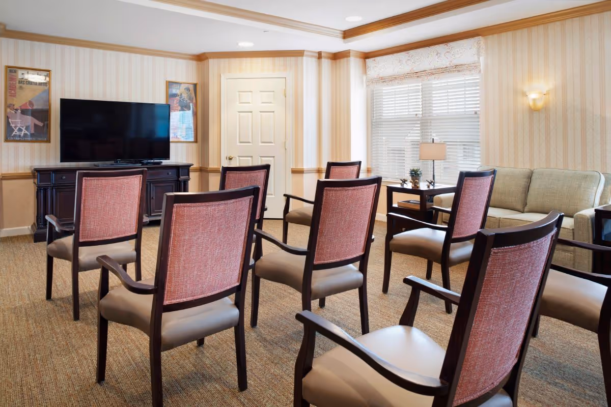 Communal sitting room with several chairs arranged facing a flat-screen TV, a sofa, side table, and a large window.