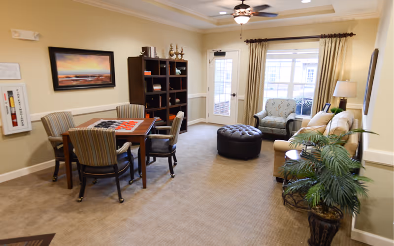 Cozy common room with a game table and chairs, bookshelf, sofa and armchair, ottoman, and a door and window to the outside.