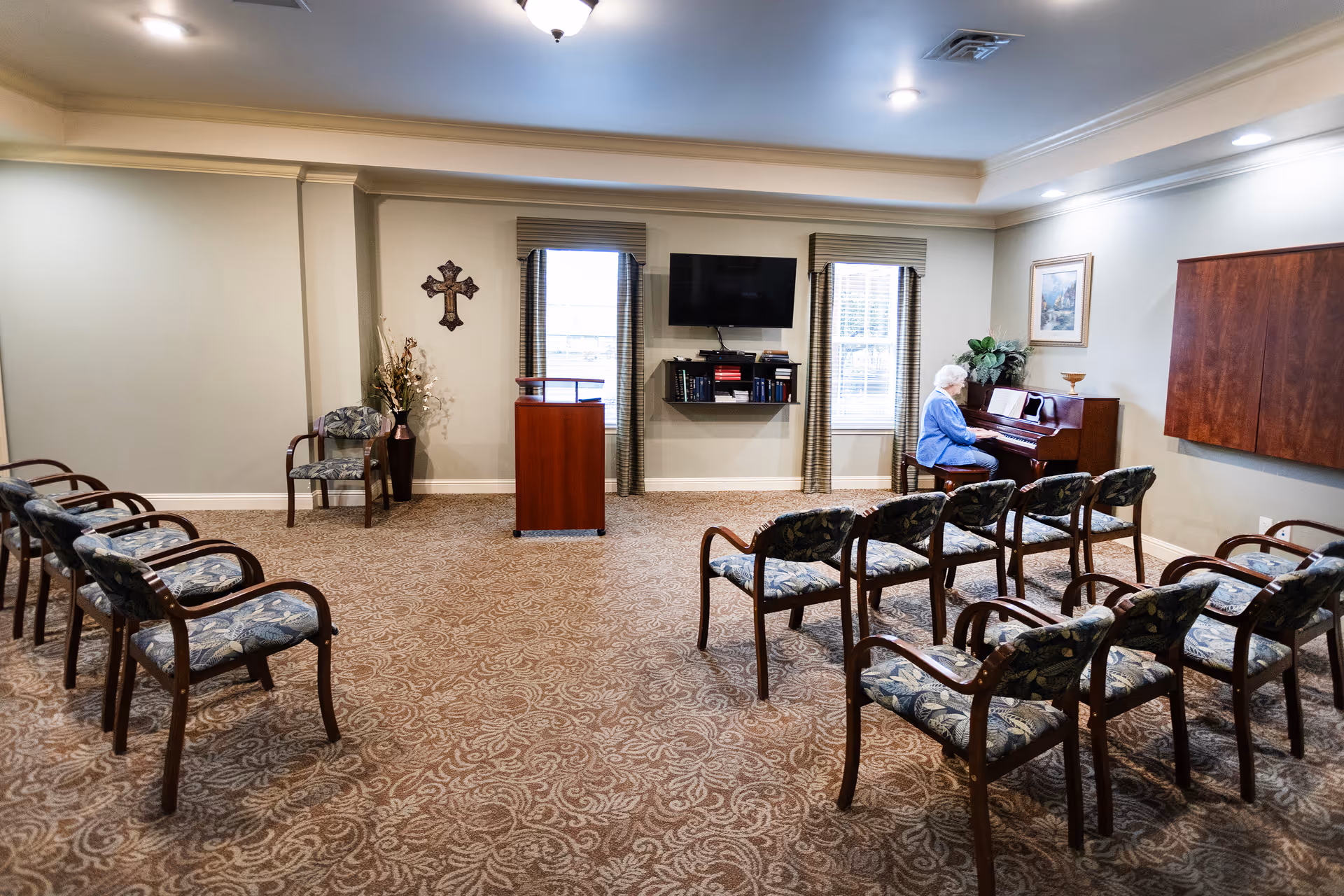 A small room with patterned carpet and several rows of chairs facing a podium and a wall-mounted TV. An elderly person is seated at a piano on the right side of the room. The room has two windows with blinds and a decorative cross on the wall.