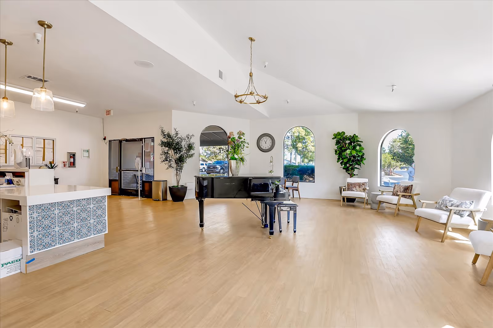 A spacious and bright common area in a senior living facility featuring a black grand piano in the center, several white armchairs with cushions arranged near large arched windows, a clock on the wall, and potted plants. The room has light wood flooring and white walls, with a reception desk on the left side decorated with blue patterned tiles.