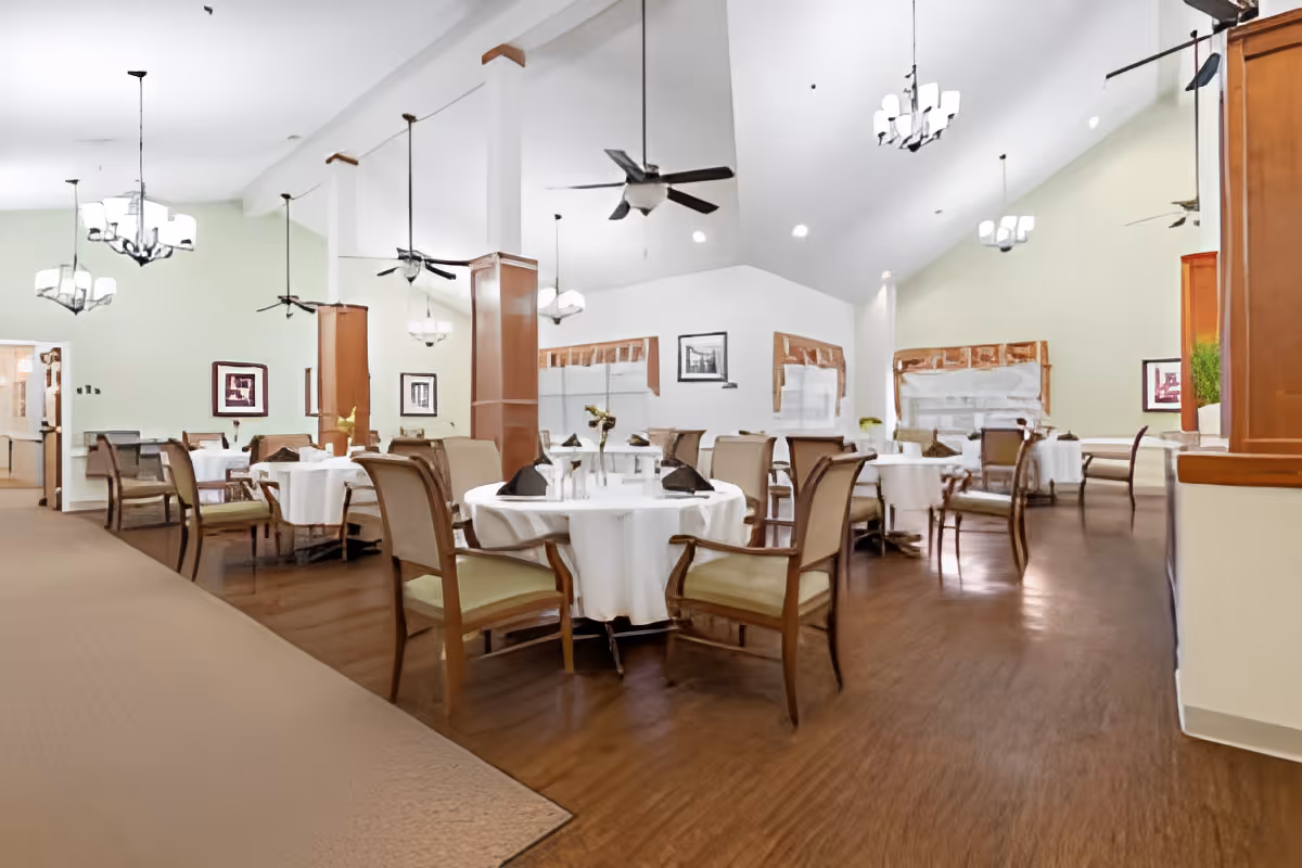 A spacious dining room with round tables covered in white tablecloths, each set with napkins and small flower arrangements. The room features wooden chairs with green cushions, high vaulted ceilings with ceiling fans, and multiple hanging light fixtures. The walls are painted light green and white, and framed artwork is visible on the walls.