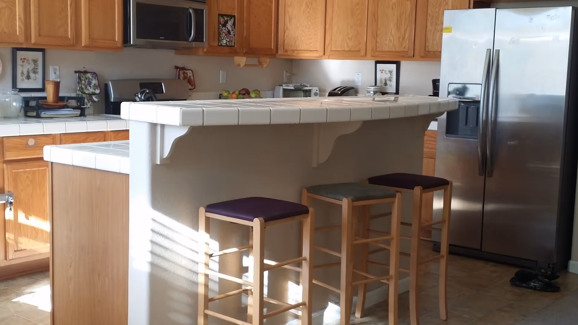 A kitchen with wooden cabinets, a white tiled countertop island with three wooden bar stools, and a stainless steel refrigerator. Various kitchen appliances and decorative items are on the countertops.