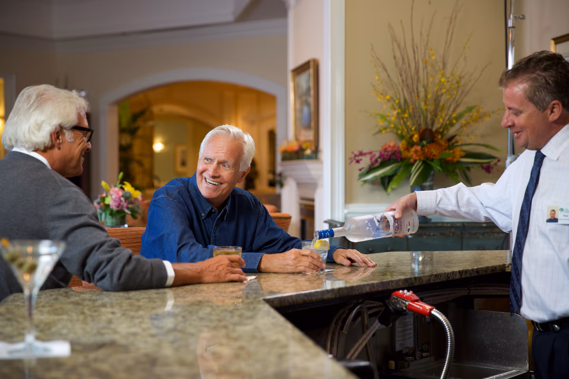 Two elderly men sitting at a marble countertop bar, smiling and holding drinks, while a bartender pours a drink into a glass. The setting is a warmly lit indoor area with floral arrangements and framed artwork on the walls.