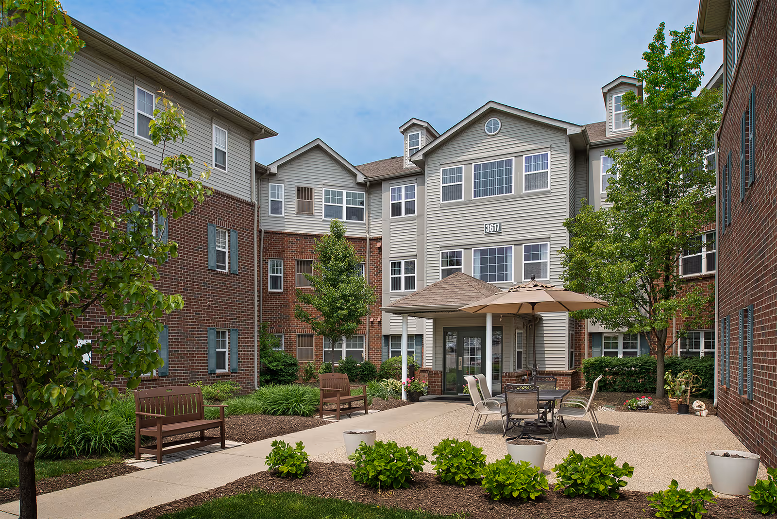 Outdoor courtyard area of a senior living facility with benches, a table with chairs and an umbrella, surrounded by trees and shrubs. The building has a combination of brick and siding exterior with multiple windows and a small covered entrance.