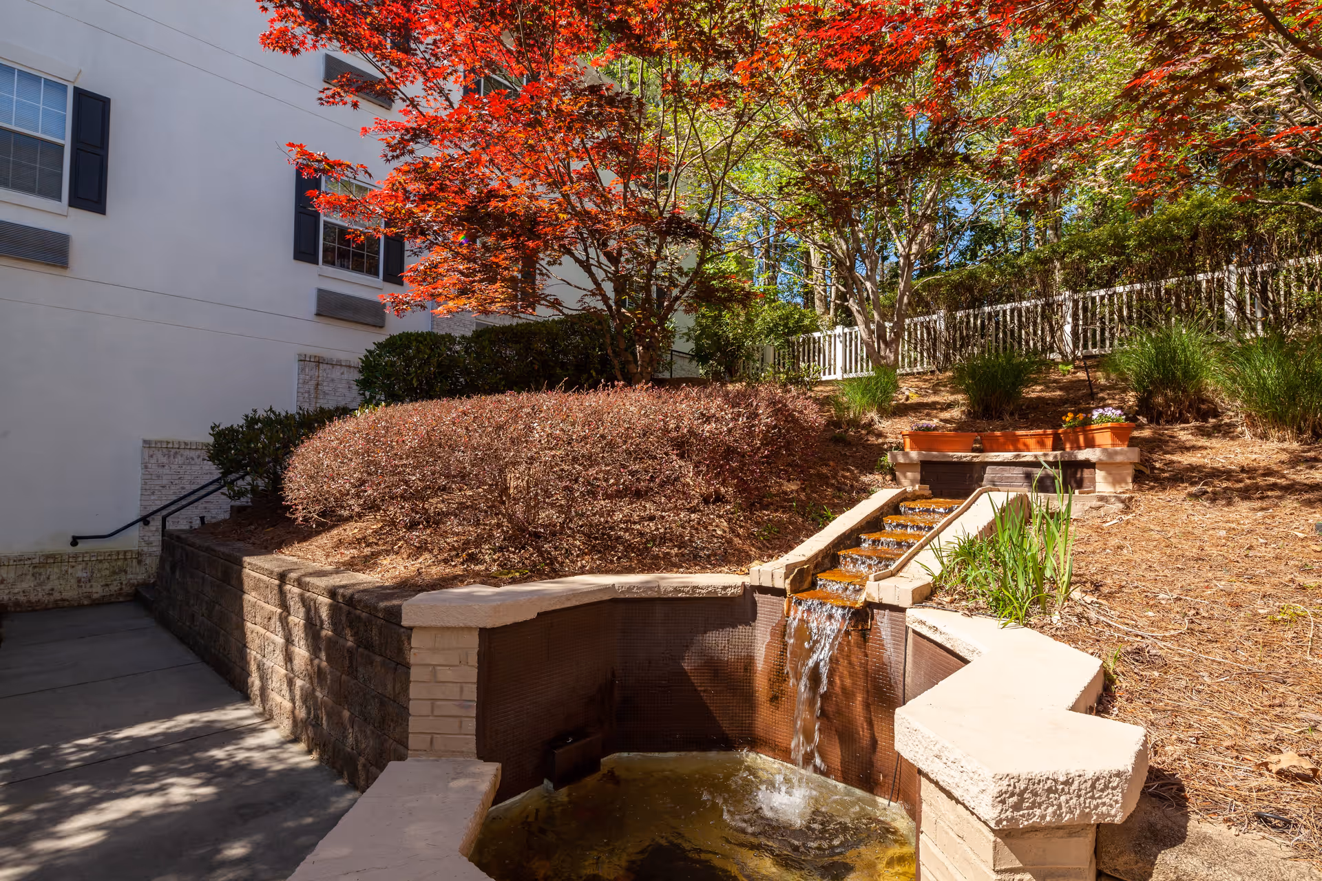 Outdoor garden area with a small cascading water feature surrounded by stone walls and landscaping. There are bushes, a tree with red leaves, and other greenery. A white building with windows and black shutters is visible on the left side, and a white fence is in the background.