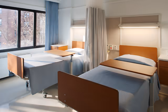 A bright hospital or nursing facility room with two beds separated by a privacy curtain. Each bed has a wooden headboard and footboard, a light blue bedspread, and an adjustable overbed table. There is a large window with blue curtains letting in natural light, and bedside cabinets next to each bed.