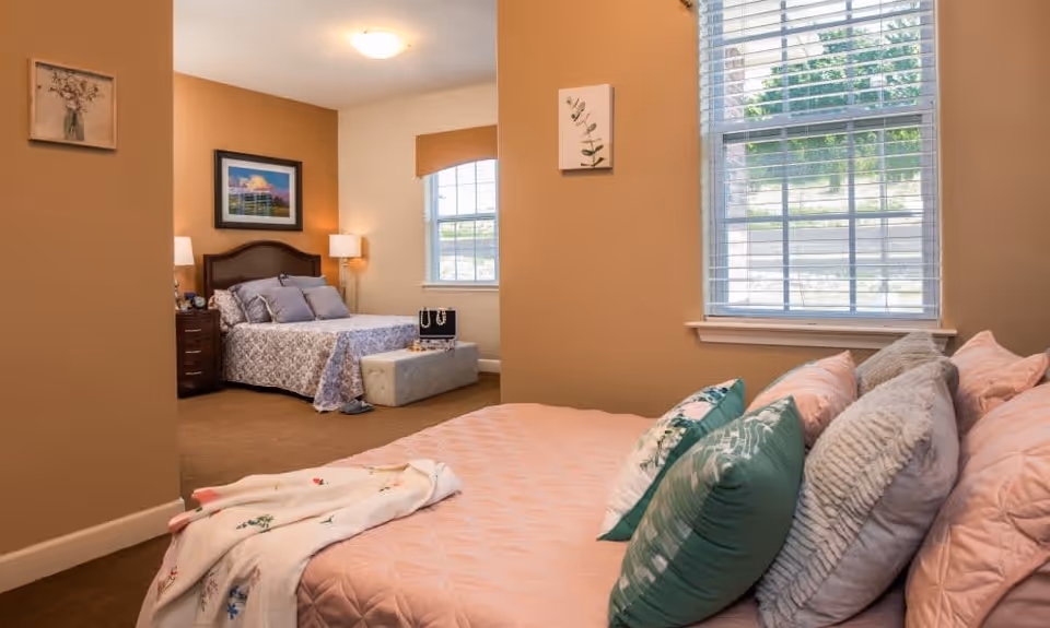 Interior view of a senior living facility bedroom with two beds. The foreground shows a bed with pink bedding and multiple pillows, next to a window with blinds. In the background, there is another bed with patterned bedding, a nightstand with a lamp, and framed artwork on the walls. The room has beige walls and carpeted floors.
