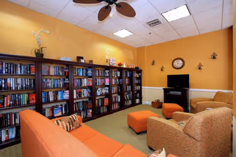 A cozy lounge with tall bookshelves along one wall, orange sofas and armchairs, a TV, and a ceiling fan.