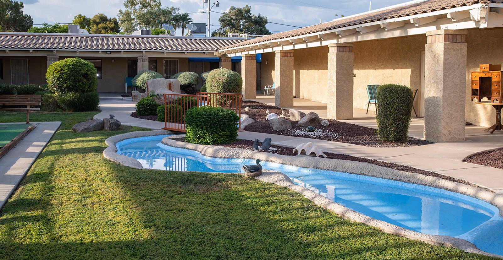 Outdoor courtyard area at Madison Meadows Retirement Community featuring a small decorative water feature with blue water, manicured bushes, a green lawn, a bocce ball court, and a covered walkway with columns and chairs.