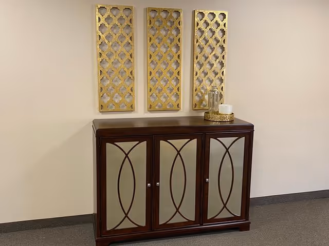 A dark wooden cabinet with four mirrored doors featuring decorative curved designs, placed against a beige wall. Above the cabinet are three vertical gold decorative panels with a lattice pattern. On top of the cabinet is a gold tray holding a glass jar and a white candle.