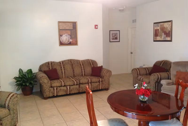 A tiled common living room with patterned sofas and armchairs, a round wooden table with a vase of red flowers, wall art, and a potted plant.