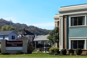 Exterior view of CareCore At Westmoreland facility showing a green and beige building with large windows, a sign reading 'Westmoreland Place' on a lawn, and a backdrop of trees and hills under a clear blue sky.