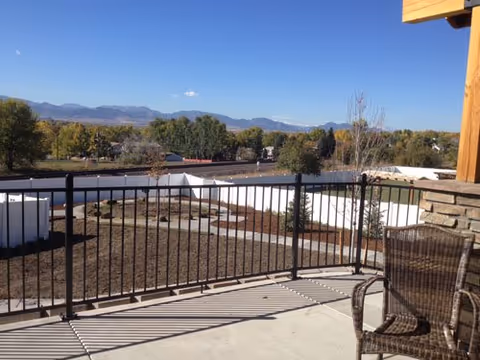 View from a balcony or patio area with a black metal railing, a wicker chair on the right, and a landscaped yard with white walls and small trees. In the background, there are trees and mountains under a clear blue sky.