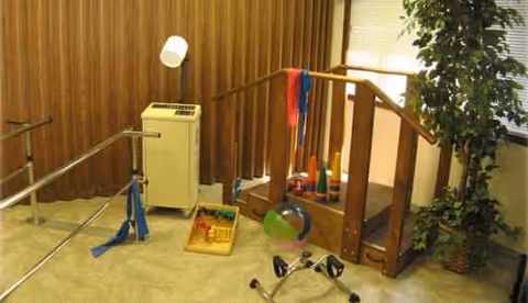 Interior rehab room with parallel bars, exercise bands, and scattered therapy equipment.