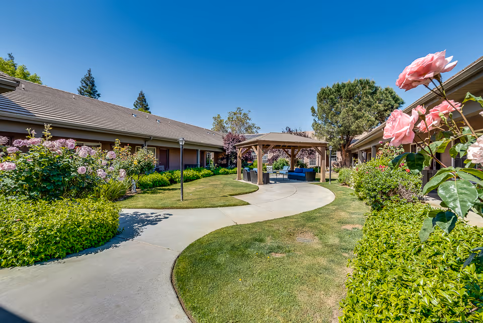 Sunny landscaped courtyard with a central wooden gazebo, curved concrete walkway, flowering bushes and single-story buildings.