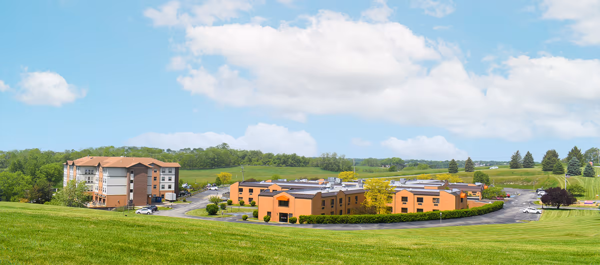 Panoramic view of William Penn Care Center showing multiple buildings with orange brick exteriors surrounded by green lawns and trees under a partly cloudy blue sky.